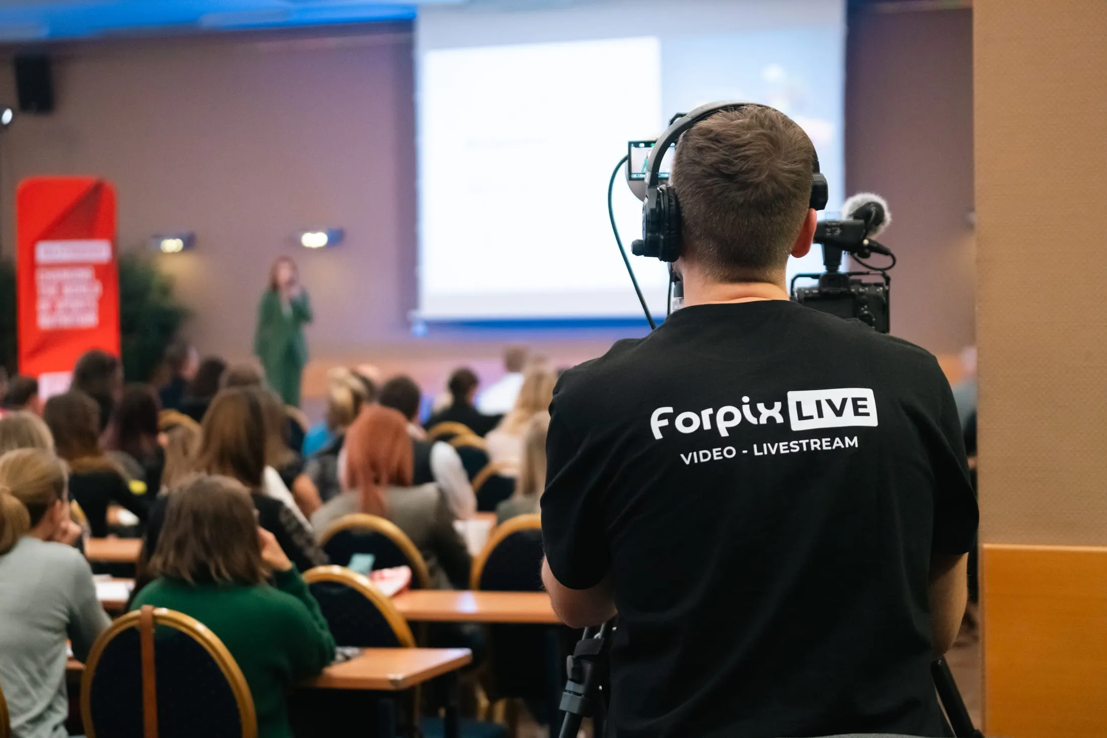 Kameraman v triku Forpix Live snímá zezadu zaplněný přednáškový sál