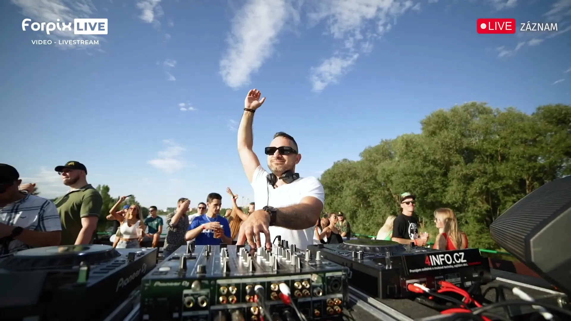 Ein DJ mit erhobener Hand hinter der Booth an Deck, um ihn herum tanzende Gäste