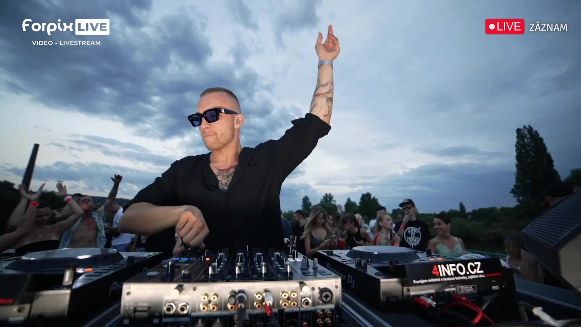 Ein DJ hinter der Booth an Deck vor dunkler werdendem Himmel, die Menge um ihn herum mit erhobenen Händen