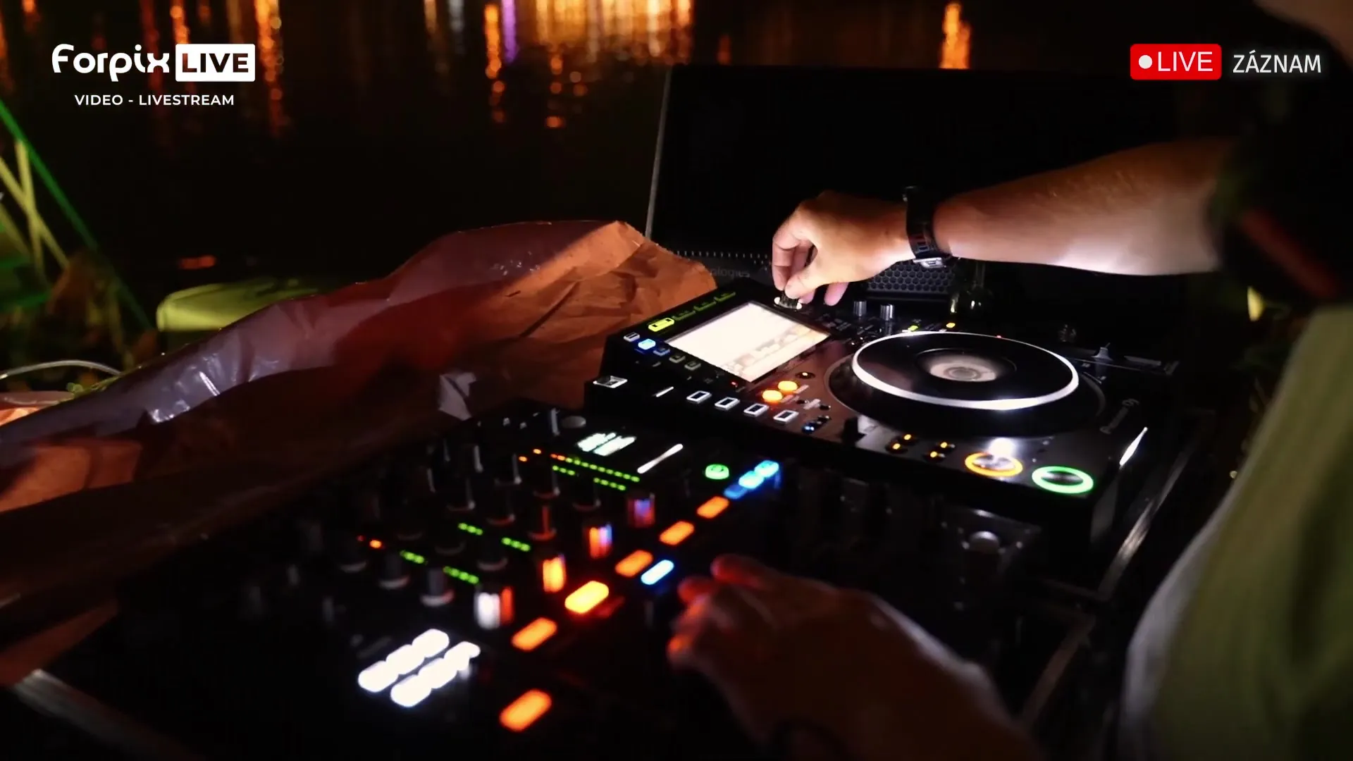 Die Hände eines DJs an den Decks bei Nacht, dahinter Lichter auf der Oberfläche der Moldau