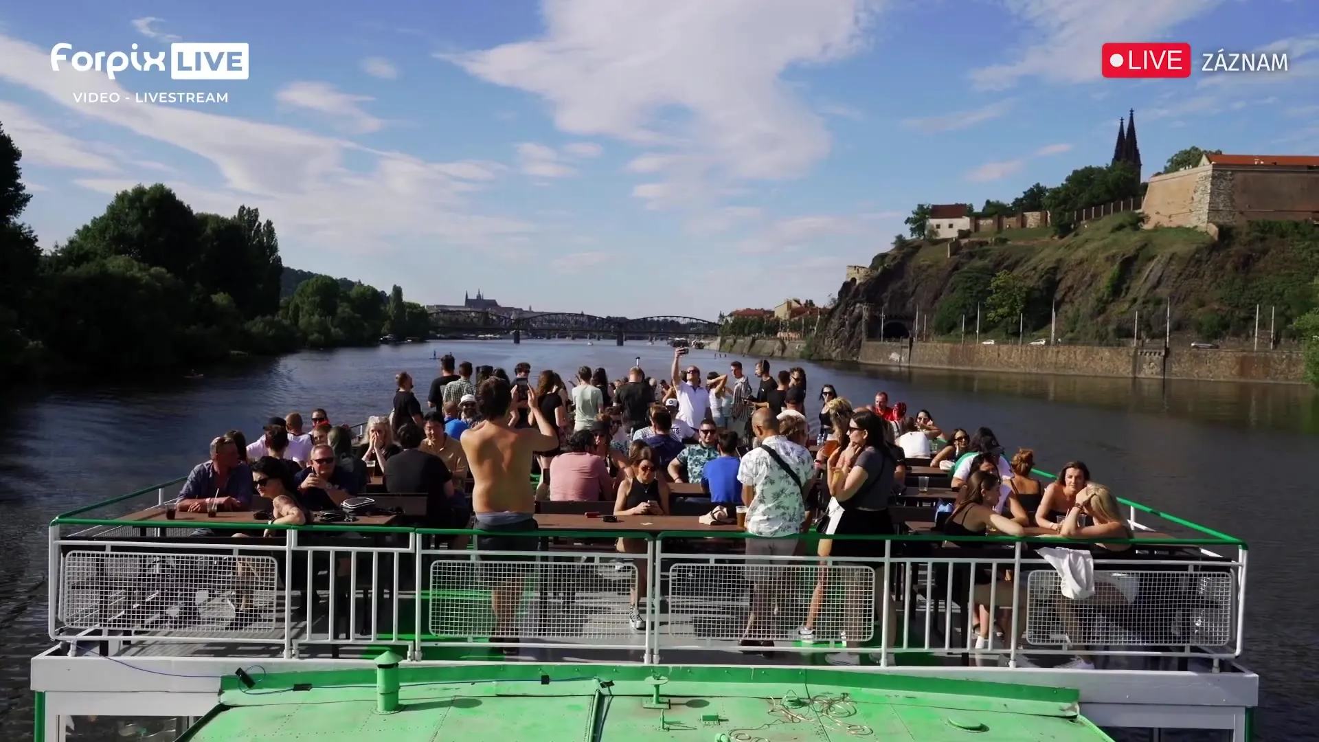 Das volle Oberdeck eines Fahrgastschiffs, dahinter Vyšehrad und eine Eisenbahnbrücke über der Moldau