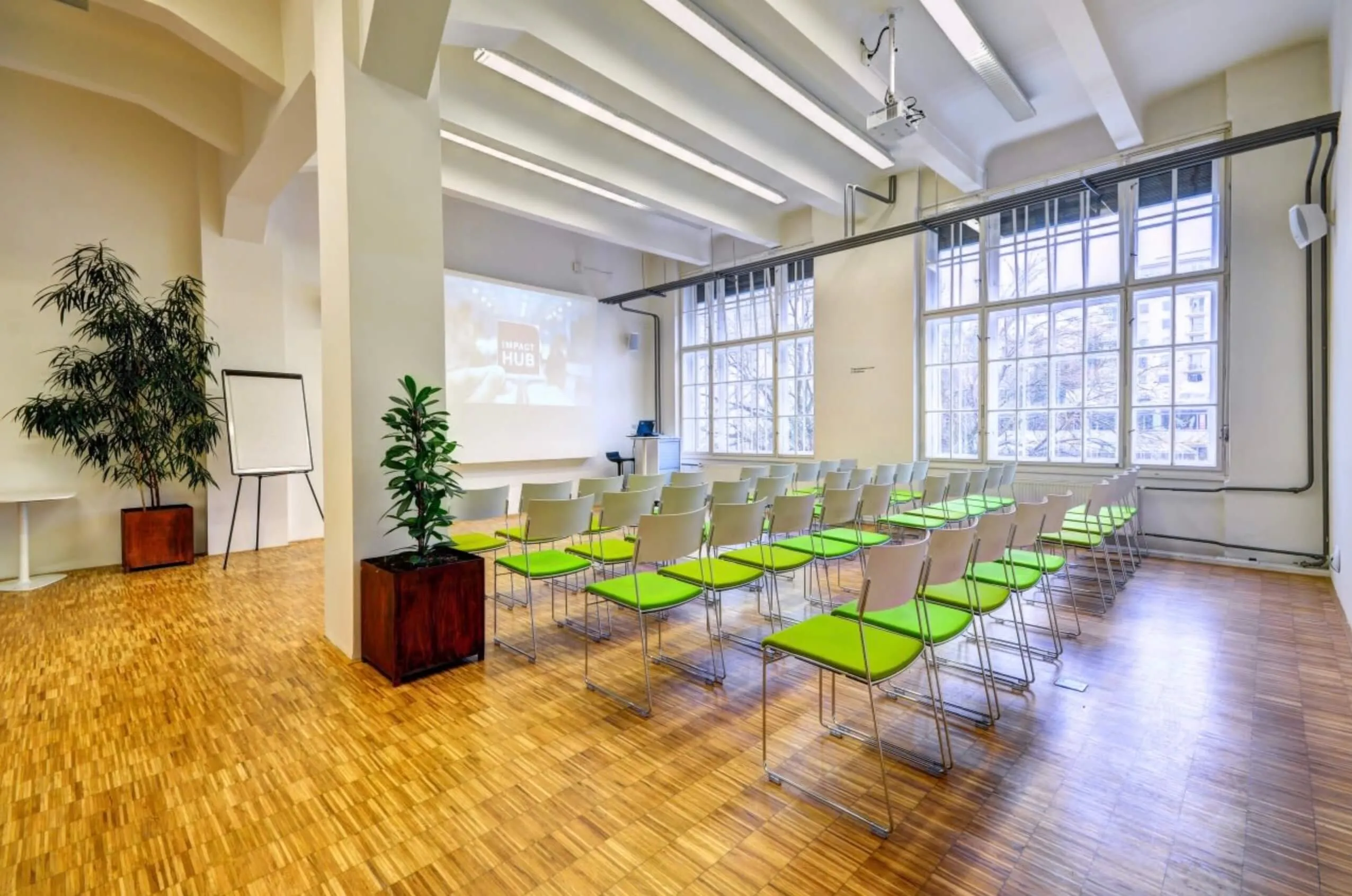 The bright Impact Hub room with parquet, tall windows and rows of green chairs facing a screen